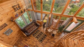 Interior d'una cabana de fusta amb un sofà, una làmpada i vista del bosc a través de grans finestres.