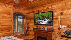 A cozy log cabin room with a fireplace, a television mounted on the wall, and a window with blinds. - Easy Access Roads*In Pigeon Forge*Jacuzzi and hot tub (PIGEON FORGE)