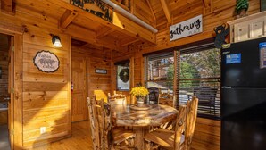 Interior de una cabaña con una mesa de comedor de madera, sillas, un refrigerador y un letrero que dice 'The Gathering Place'.