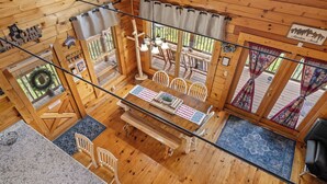 A wooden cabin with a dining area featuring a table with an American flag placemat, white chairs, and a wooden bench. There are two windows with curtains, a wooden ceiling, and a sign that reads "Adventure Awaits."