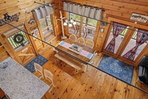Uma cabana de madeira com área de jantar, incluindo uma mesa com jogo americano da bandeira dos EUA, cadeiras brancas e um banco de madeira. Possui duas janelas com cortinas, teto de madeira e uma placa com a inscrição 'Aventura Te Espera'.
