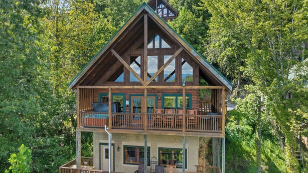 A two-story log cabin with a balcony on the upper floor, surrounded by dense forest.