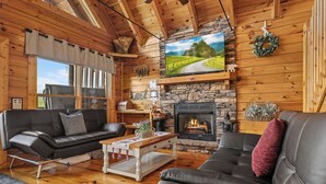 A cozy cabin interior with a stone fireplace, leather sofas, a television, and wooden walls.