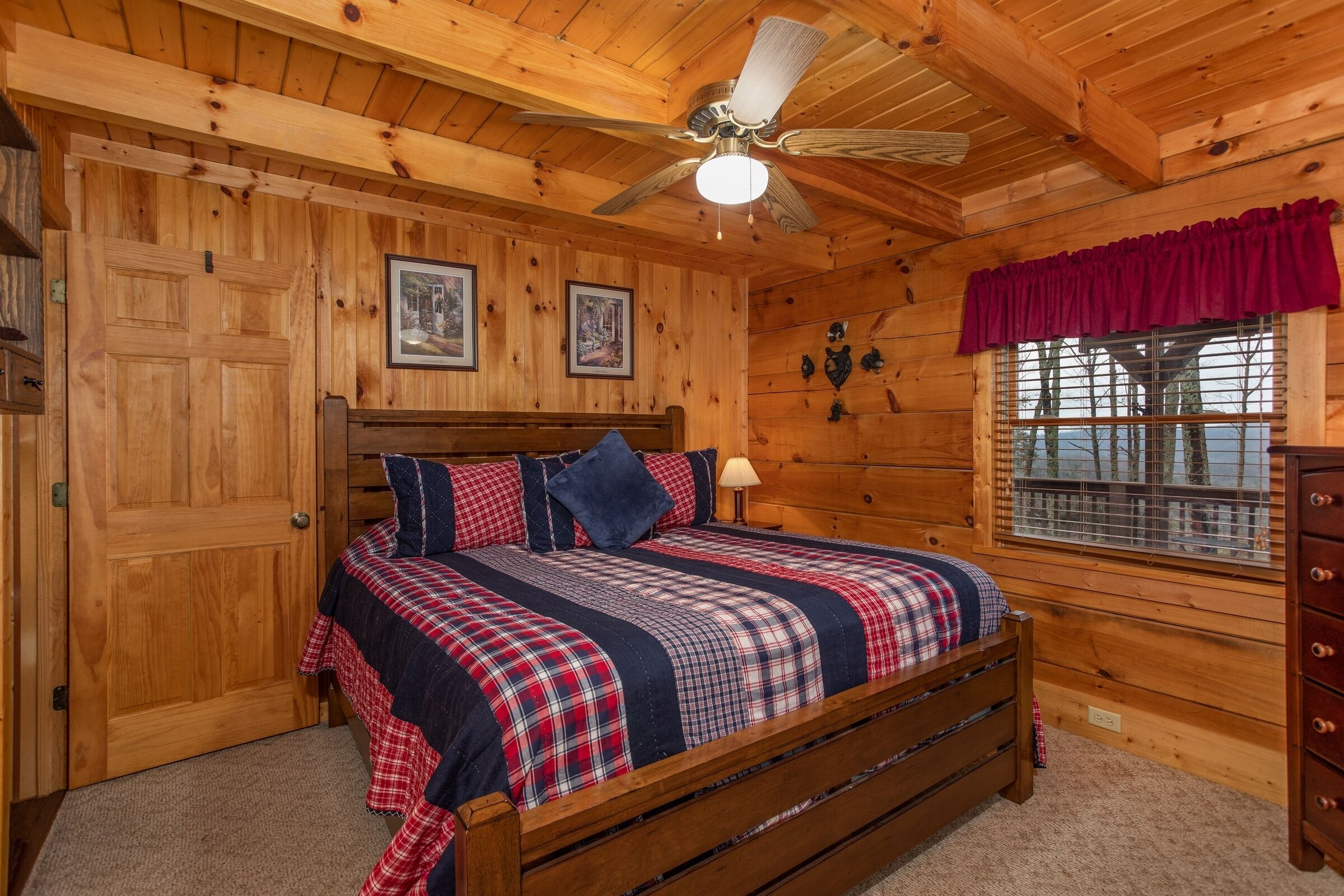 A wooden cabin bedroom with a bed, ceiling fan, wooden walls, and a window with a view.