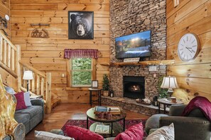 Interior de una cabaña de troncos con chimenea de piedra, paredes de madera, televisor y un reloj.