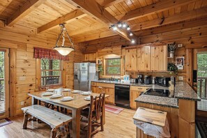 A wooden cabin interior with a dining table, chairs, a kitchen with granite countertops, and a refrigerator.