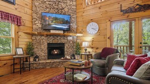 A cozy cabin interior with a stone fireplace, a television mounted on the wall, a clock, and a comfortable seating area with a red rug. - Resort*Luxury*Close to Pigeon Forge*Game Loft*Seasonal Pool (SEVIERVILLE)