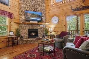 A cozy cabin interior with a stone fireplace, a television mounted on the wall, a clock, and a comfortable seating area with a red rug.