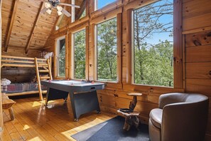 A wooden cabin with a pool table, a loft bed, and large windows offering a view of trees.