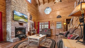 A cozy log cabin interior with a stone fireplace, wooden walls, a large window, and a comfortable seating area. - Pigeon Forge*Mountain Views*Family Fun*Game Room*Hot Tub (PIGEON FORGE)