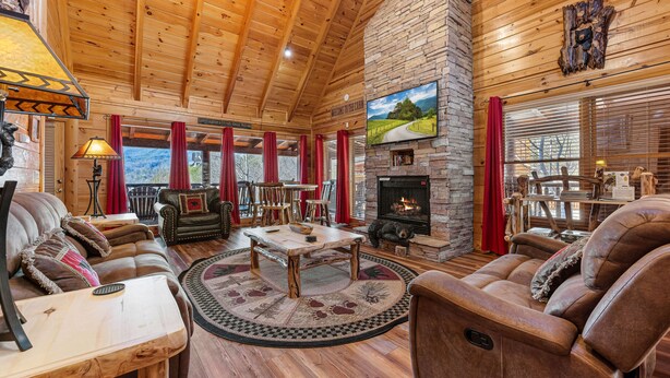 A cozy log cabin interior with a stone fireplace, leather sofas, a wooden dining table, and a large TV mounted on the wall. - Pigeon Forge*Mountain Views*Family Fun*Game Room*Hot Tub (PIGEON FORGE)
