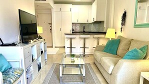 A modern living room with a glass coffee table, a sofa with teal cushions, a TV on a white cabinet, and a kitchen area in the background.
