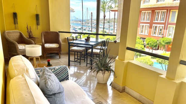 A balcony with wicker furniture, a dining table, and a view of a marina and buildings.