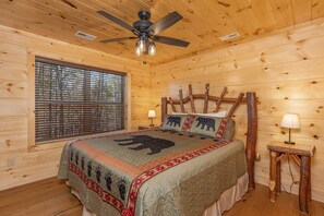 Une chambre dans une cabane en bois, avec un lit, un ventilateur de plafond, une fenêtre avec des stores et une petite table avec une lampe.
