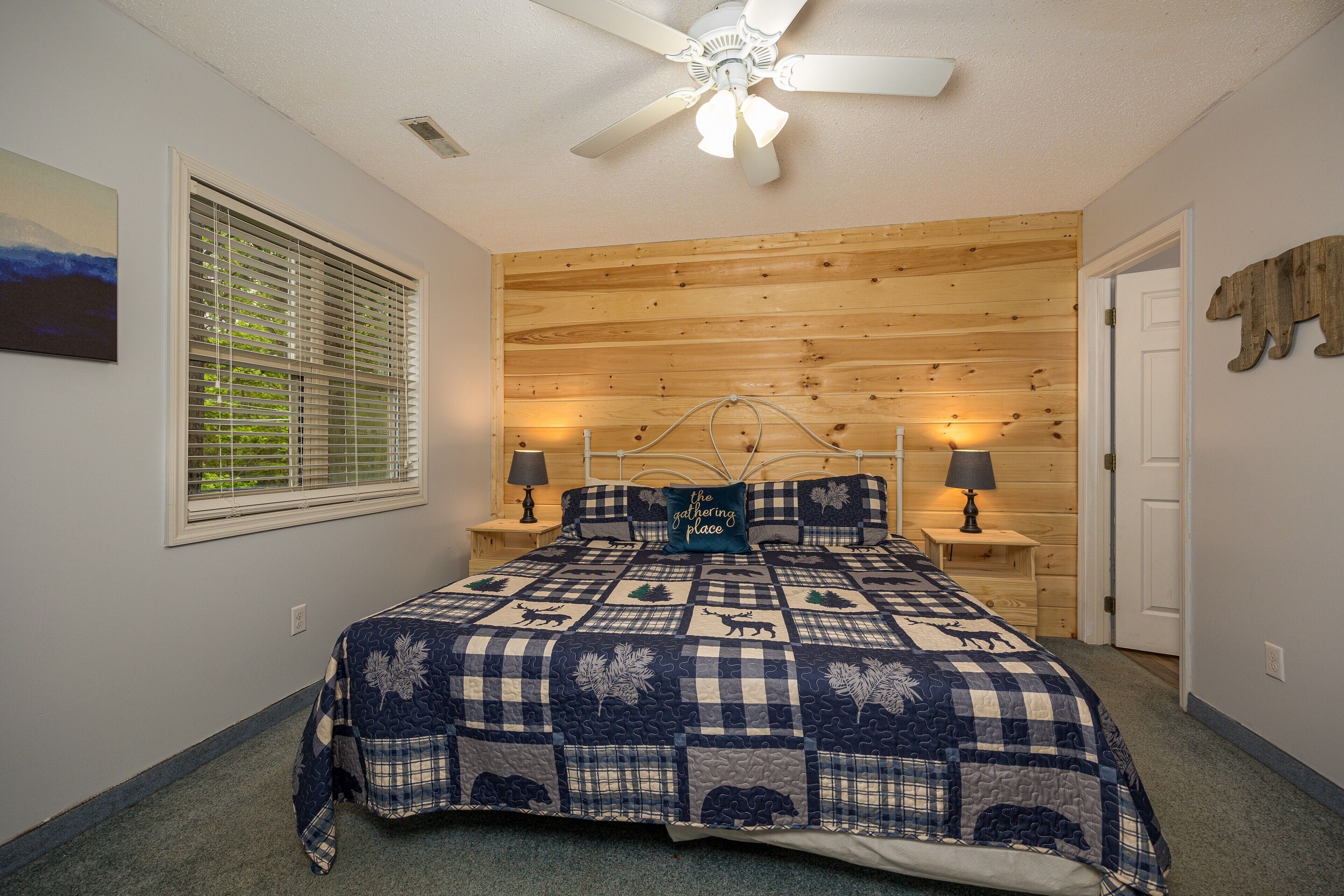 A bedroom with a wooden ceiling, a bed with a blue and white quilt, two bedside tables with lamps, a window with blinds, and a door.