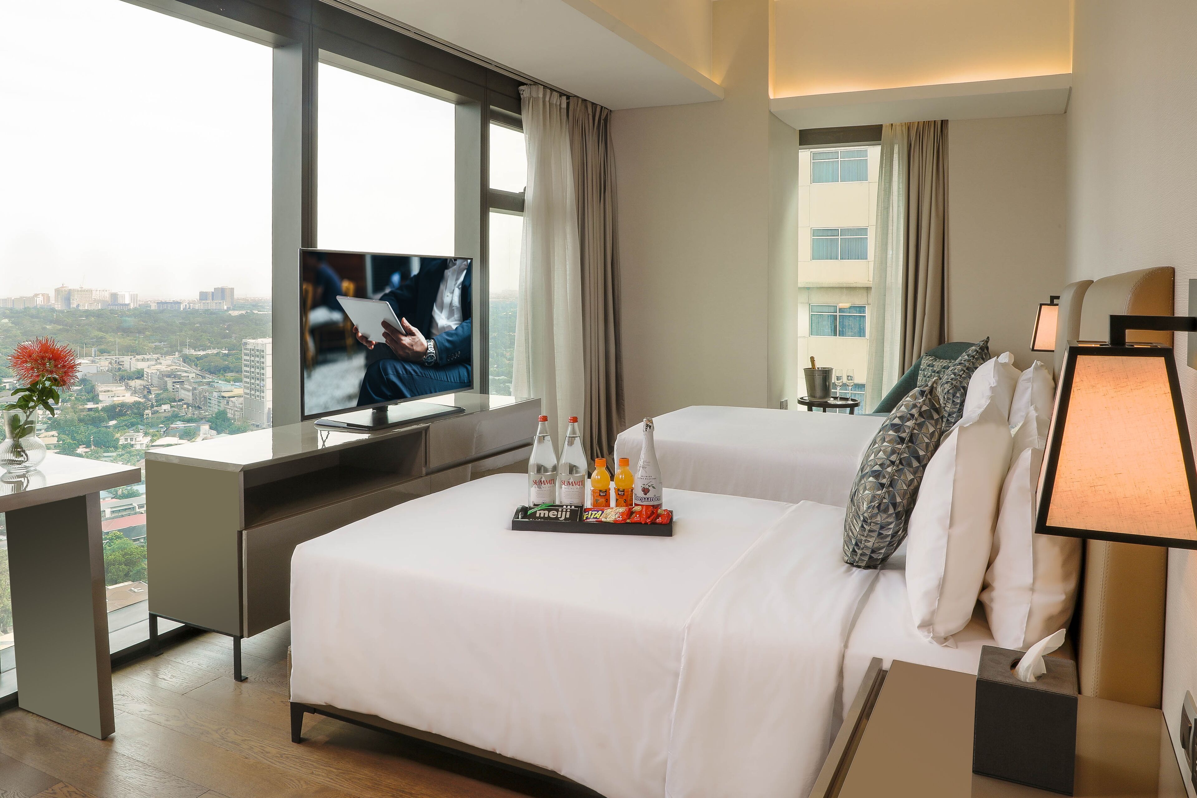 A hotel room with two beds, a desk, a TV, and a city view.