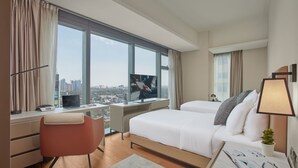 Une chambre d’hôtel avec une grande fenêtre, un bureau avec un ordinateur portable, un lit avec des oreillers et une petite plante.