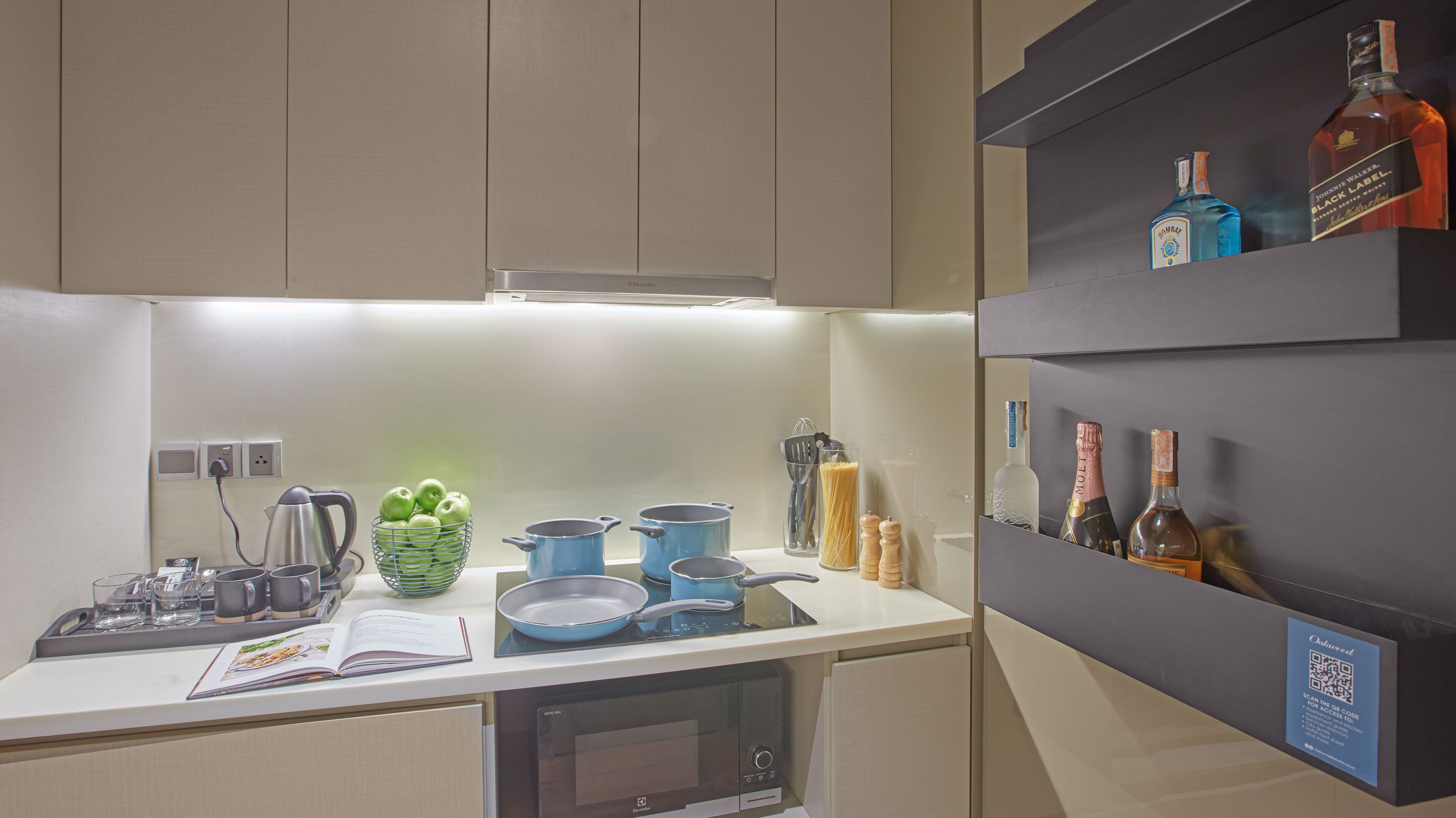 A modern kitchen with a built-in oven, a microwave, and a coffee machine. There are several pots and pans on the counter, and a basket of green apples. A shelf above the counter holds various bottles of alcohol.