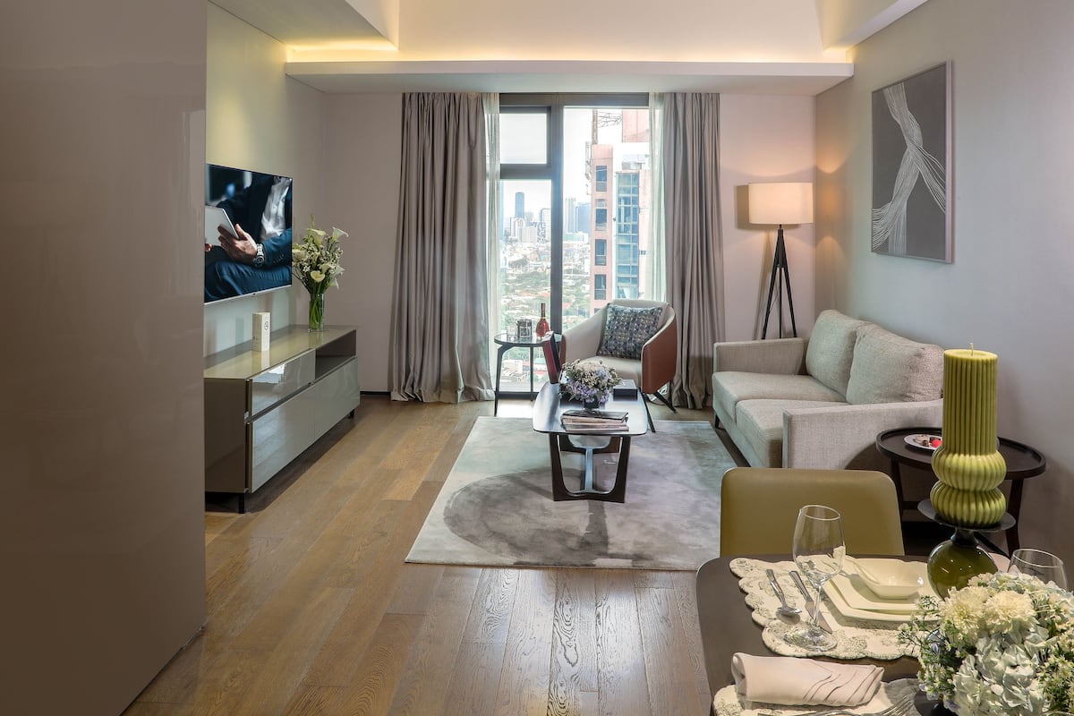 Une chambre d’hôtel moderne dotée d’une grande fenêtre, d’un canapé, d’une table à manger dressée pour deux personnes et d’un téléviseur fixé au mur.