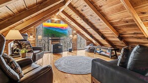 Un accogliente soggiorno mansardato con travi in legno, una grande TV, divani in pelle e un camino.