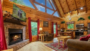 A log cabin interior with a fireplace, wooden furniture, a kitchen, and large windows. - Gatlinburg*Hot Tub*Wifi/Cable*Resort Pool (Seasonal) (GATLINBURG)