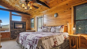 A wooden cabin bedroom with a bed, a stone fireplace, a television, and a window with blinds.