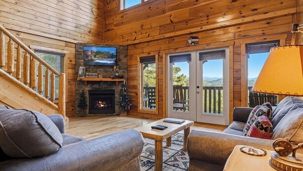 A log cabin interior with a fireplace, a television, a sofa, and a view of the outdoors.