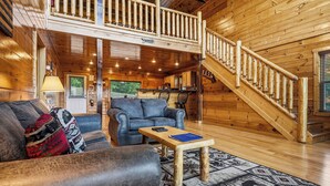A wooden cabin interior with a living room, a dining area, and a staircase leading to a loft.