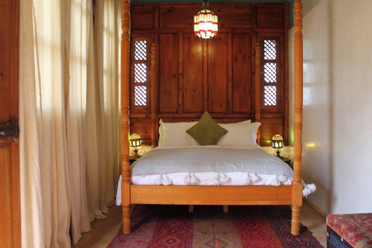 A wooden four-poster bed with a white bedspread and tassels, two bedside lamps, and a patterned rug.