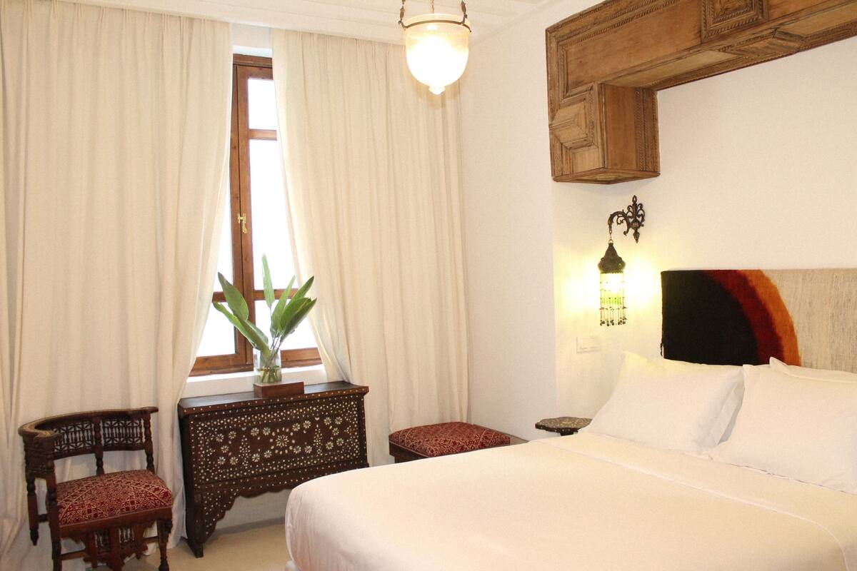 A bedroom with a large bed, a wooden headboard, a bedside table with a lamp, a chair, and a window with curtains.
