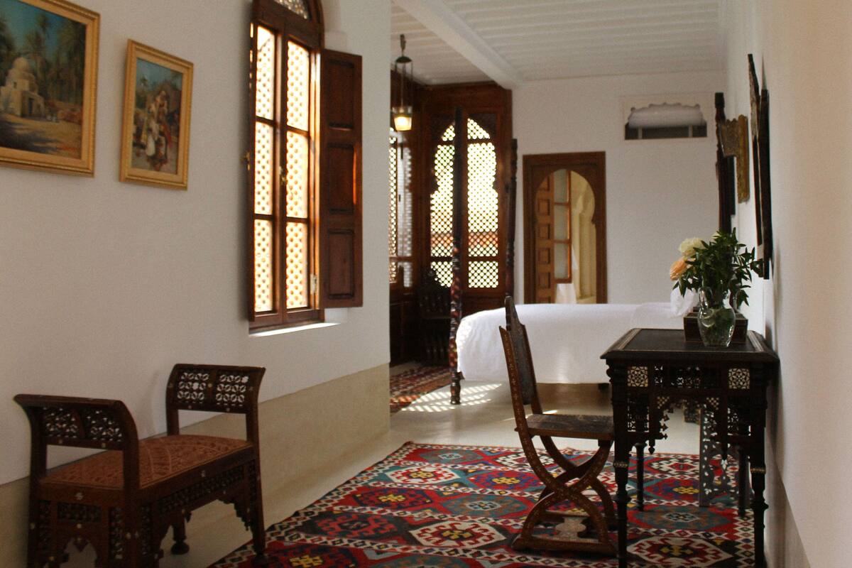 A room with a bed, a chair, a table with a vase, and a patterned rug.