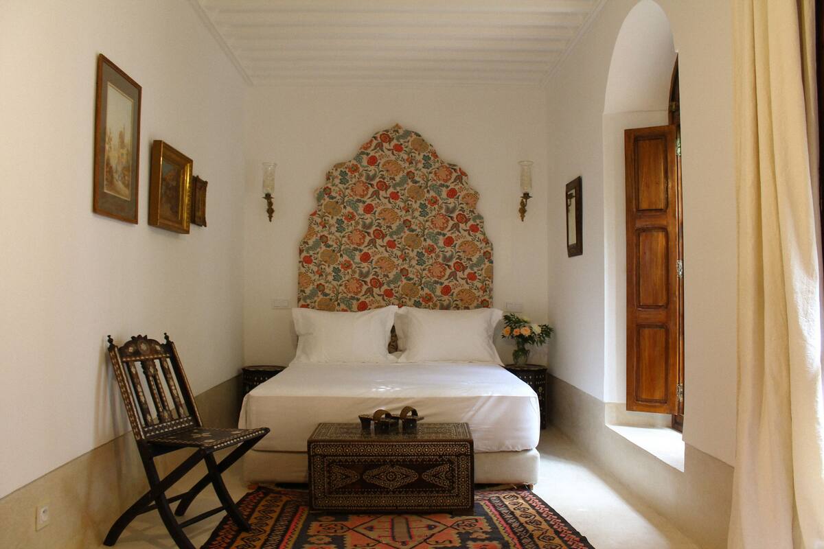 A bedroom with a bed, a chair, a nightstand, a decorative headboard, and a window with curtains.