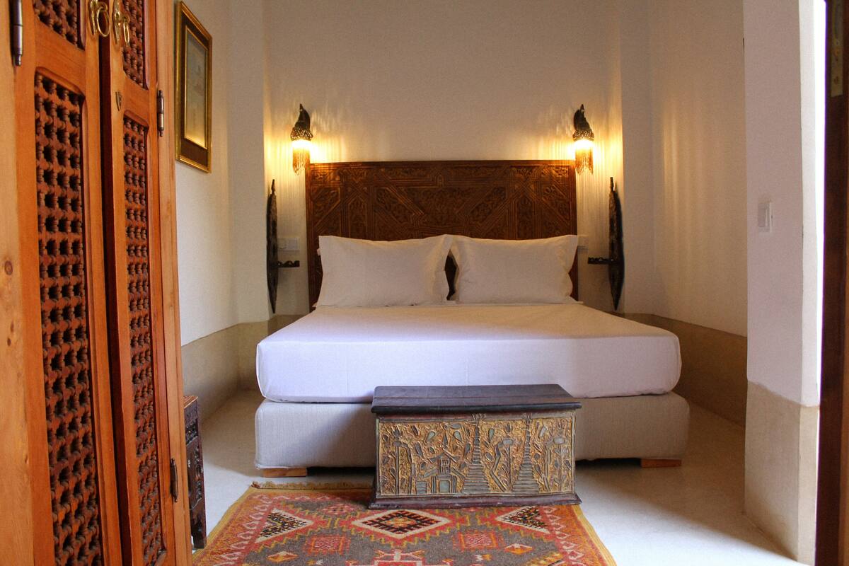 A bedroom with a large bed, two wall-mounted lamps, a wooden headboard, and a patterned rug.