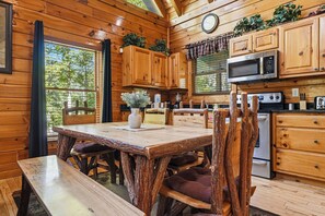 A wooden cabin interior with a dining table, chairs, a kitchen with appliances, and a window with curtains. - Gatlinburg*Resort*Cable/Wifi*Hot Tub*Seasonal Pool Access (Gatlinburg)