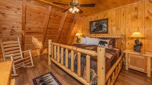 A cozy log cabin bedroom with a wooden bed, a rocking chair, a ceiling fan, and a painting on the wall. - Views*Close to Dollywood*Keurig*Pool Table*Wifi*Resort Pool (PIGEON FORGE)