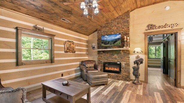 Ein gemütliches Blockhaus-Interieur mit Kamin aus Stein, Holzwänden und einem großen Fenster mit Blick auf Grünflächen.