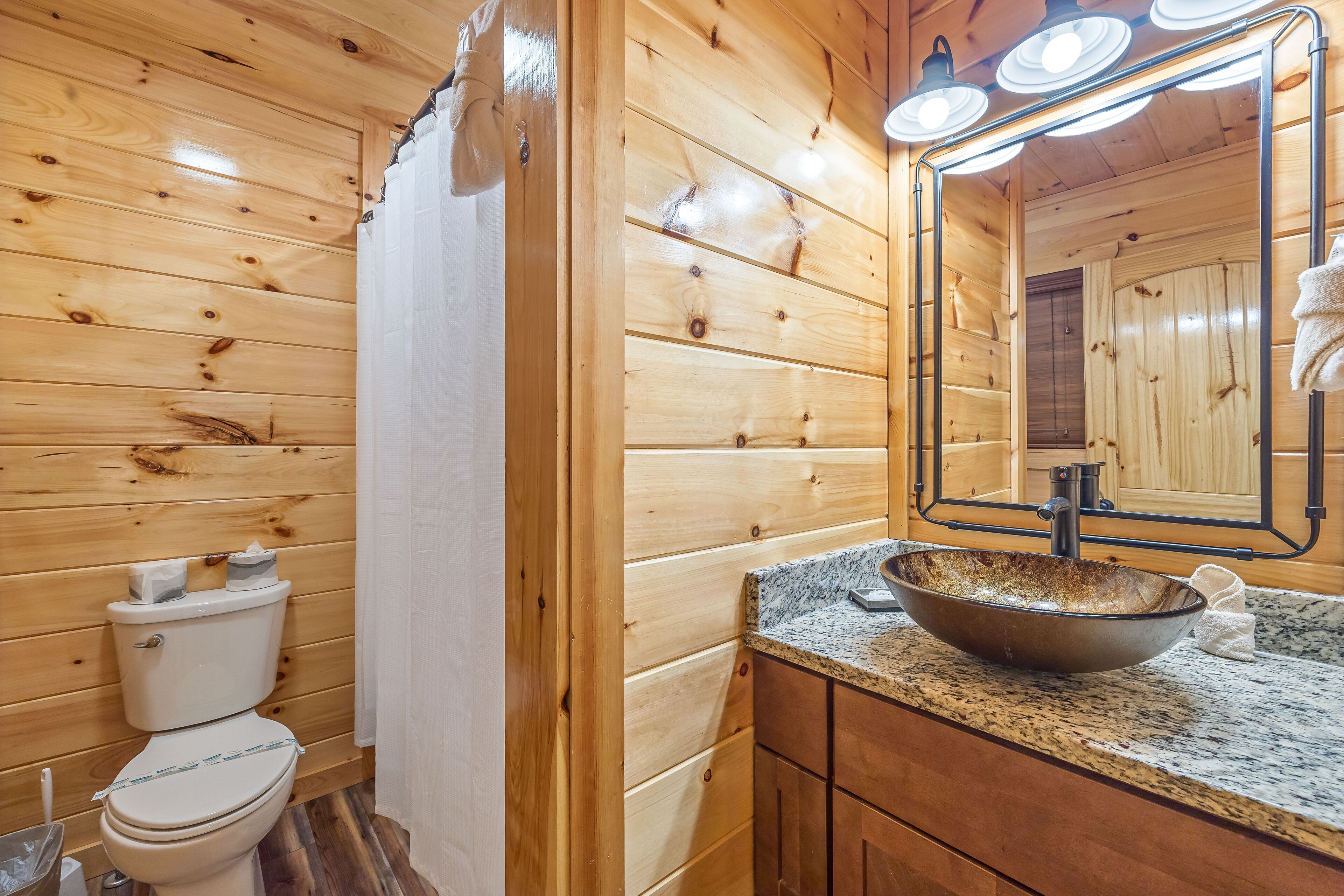 Un baño de cabaña de madera con inodoro, un espejo grande y una encimera de granito con lavabo sobre encimera.