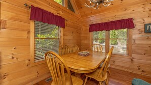 A wooden cabin interior with a dining table surrounded by chairs, a chandelier, and two windows with shutters. - Perfect Family Vacation Cabin (GATLINBURG)