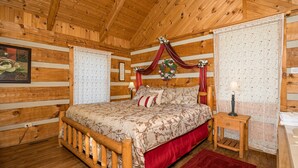 A wooden cabin bedroom with a bed, a nightstand, a red rug, and a window with curtains. - Rustic Studio*Mountain Views*Jacuzzi*Romantic (PIGEON FORGE)