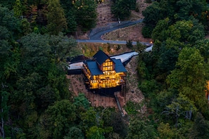 A large, multi-story house with a prominent roof and multiple windows, surrounded by dense forest.
