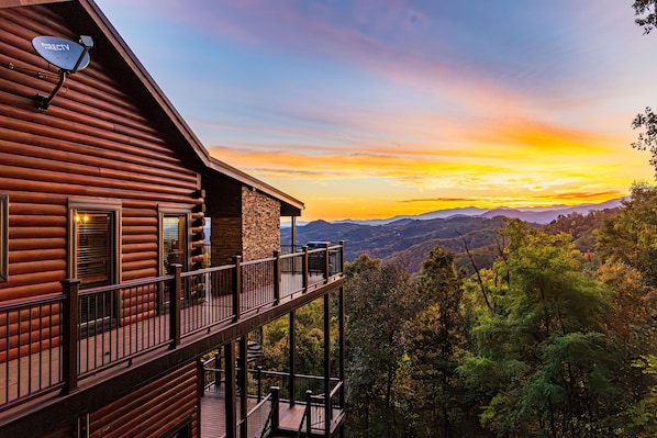 A log cabin with a balcony overlooking a forested valley and a sunset.