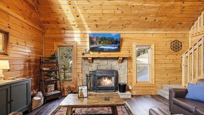 Interior acogedor de una cabaña con chimenea de piedra, paredes de madera, un televisor montado encima y una zona de estar confortable con un sofá y una mesita de centro.