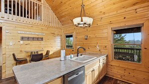 Interior de una cabaña de madera con cocina que incluye un lavamanos doble, una mesa de comedor con sillas y vistas a través de un ventanal amplio.