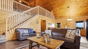 Interior de una cabaña de madera con escalera, dos sofás, una mesa de centro y una zona de cocina.