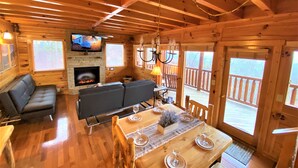 Ein gemütliches Blockhaus-Interieur mit Kamin, Holzwänden, einer Couch, einem Esstisch für vier Personen und einem an der Wand montierten Fernseher.