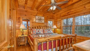 Ein Schlafzimmer in einer Blockhütte mit einem hölzernen Bett, zwei Nachttischlampen, einem Deckenventilator und großen Fenstern.