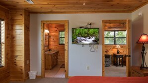 Ein Holzblockhaus-Innenraum mit an der Wand montiertem Fernseher, einem Bett mit roter Decke und einem kleinen Tisch mit Lampe.