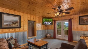 Ein gemütliches Blockhaus-Interieur mit Holzwänden, einem Deckenventilator, einem Fernseher und einem großen Fenster mit Blick ins Freie.