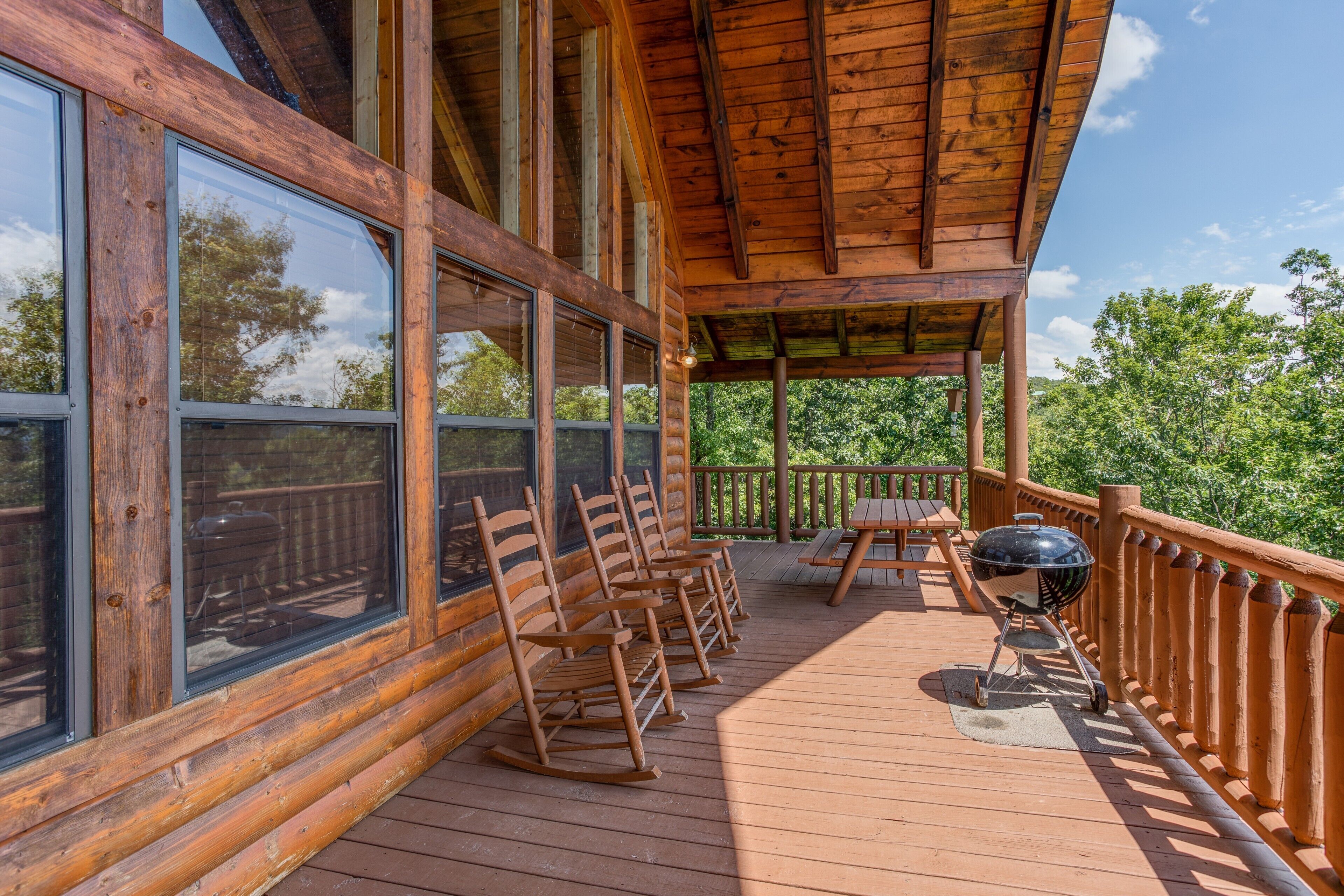 Une terrasse en bois, agrémentée de fauteuils à bascule et d’un barbecue, entourée de grandes fenêtres et d’une balustrade en bois.