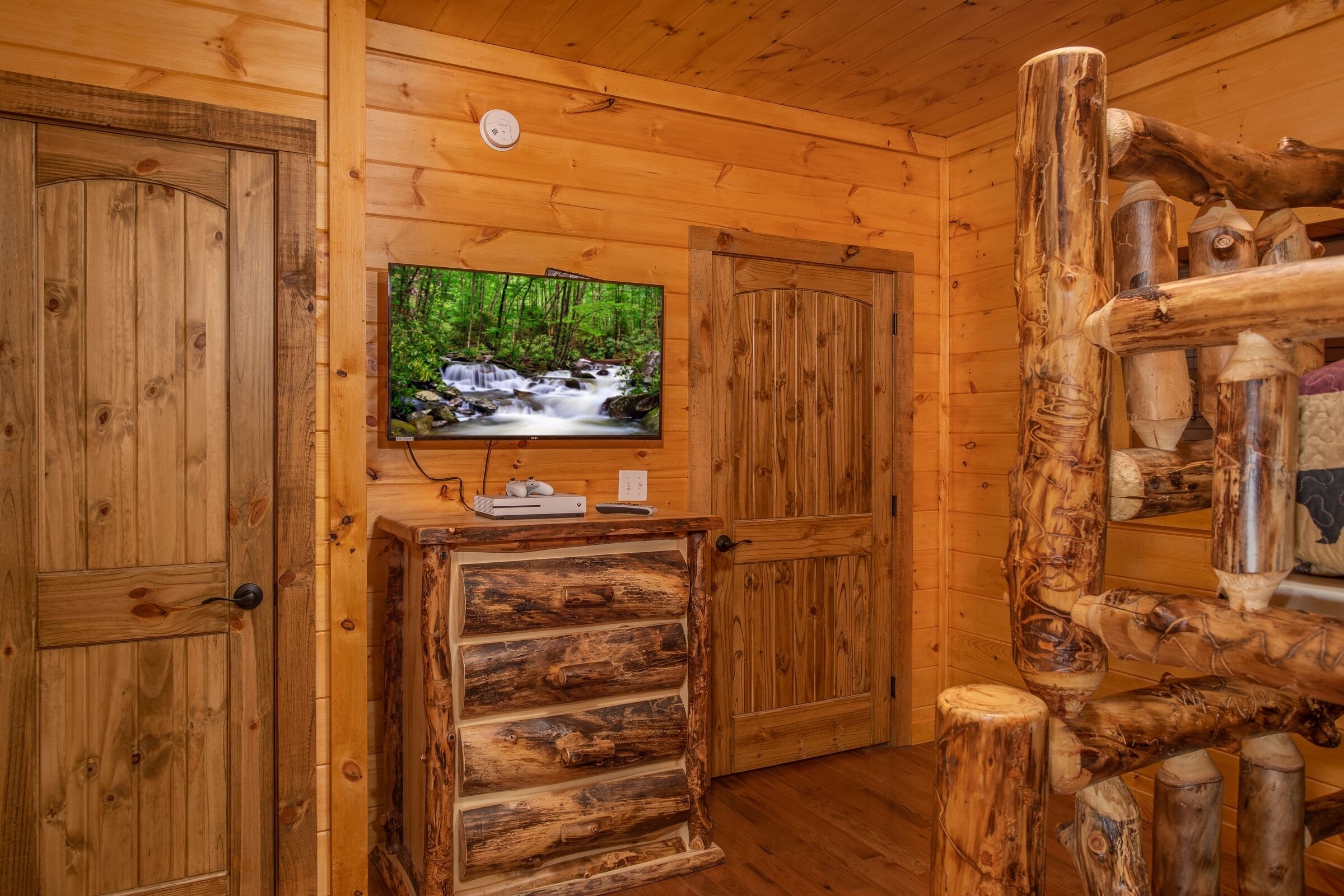 Interior de una cabaña de madera con un televisor fijado a la pared, un aparador de madera y una escalera de madera rústica.